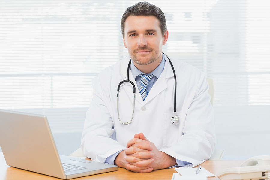 Portrait of a smiling male doctor with laptop sitting at desk in medical office