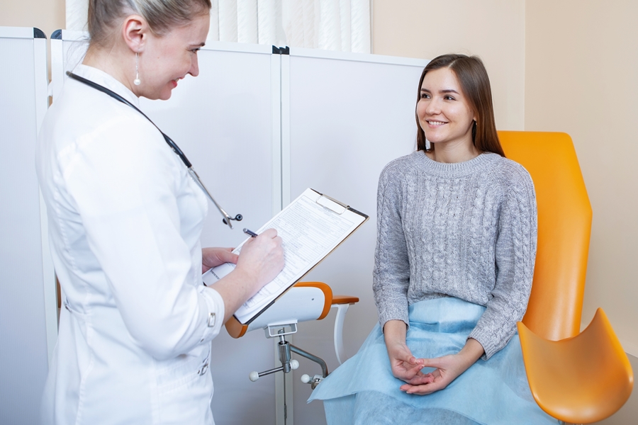 Dos mujeres, una médica de pie de costado da instrucciones a una paciente sentada en una camilla ginecologica