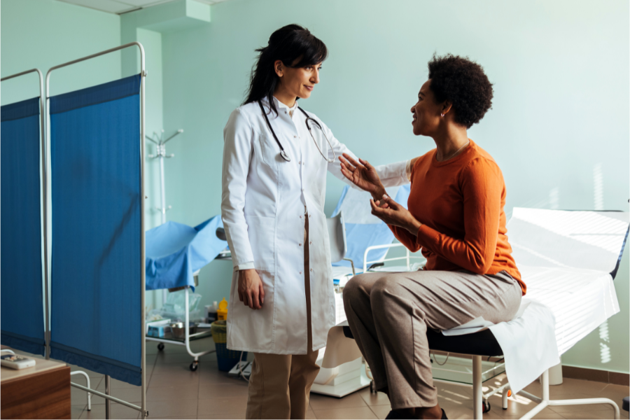 Mujer sentada sobre una camilla en una sala de examen medico hablando con una mujer medica frente a ella